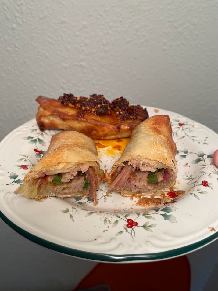 hand holding two egg rolls on a christmas themed glass plate. Egg roll in front is cut in half, showing carnitas, green onion, and cabbage inside. Egg roll in back is darker and not cut, topped with hot chili crisp.